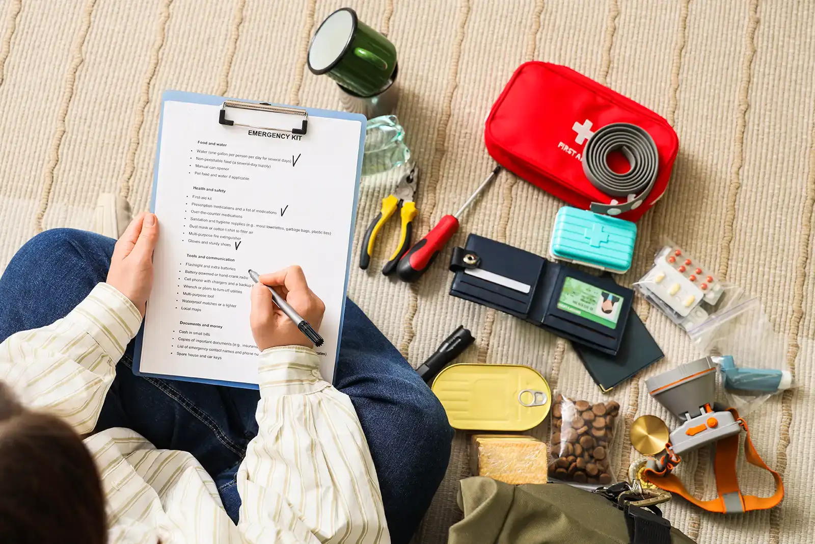 A woman sitting on the floor, marking off items on a checklist; she is surrounded by items from her emergency kit