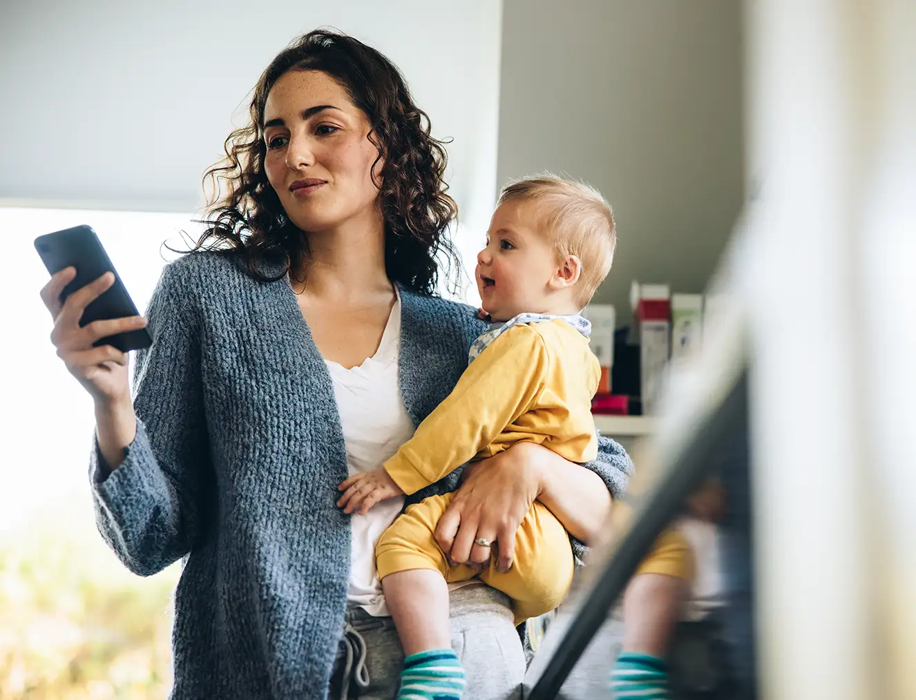 A woman holding her baby as they both smile at her phone